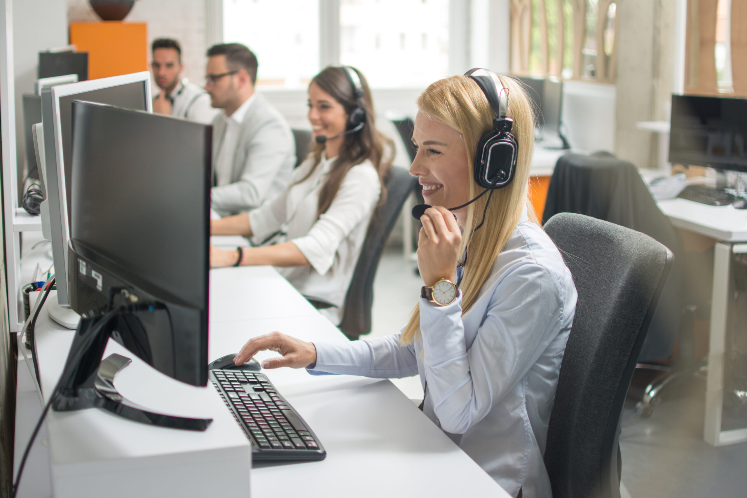 Portrait of beautiful and cheerful young woman telephone operator with headset working on desktop computer in row in customer service call support helpline business center